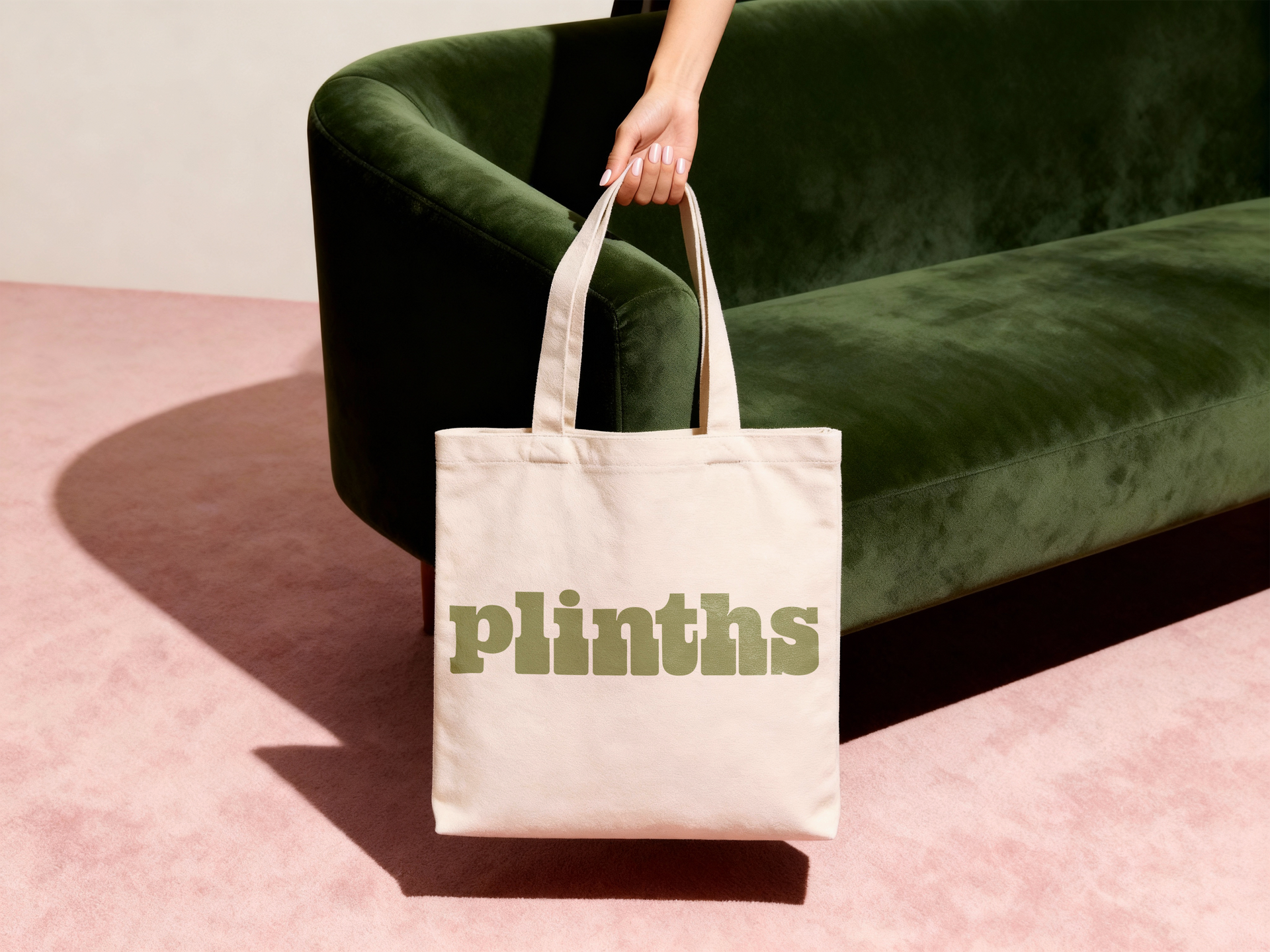 Canvas tote bag with “plinths” text, held beside a dark green sofa on a pink floor