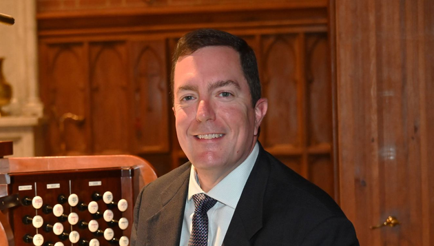 Patrick Pope in a dark suit beside a pipe organ in a wood-paneled room