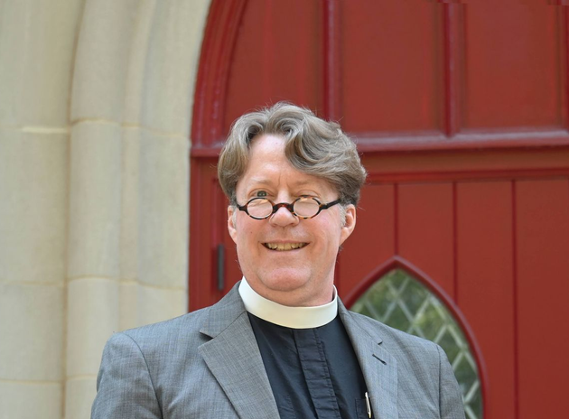 Father Brad in clerical collar and gray jacket stands smiling before a red door.