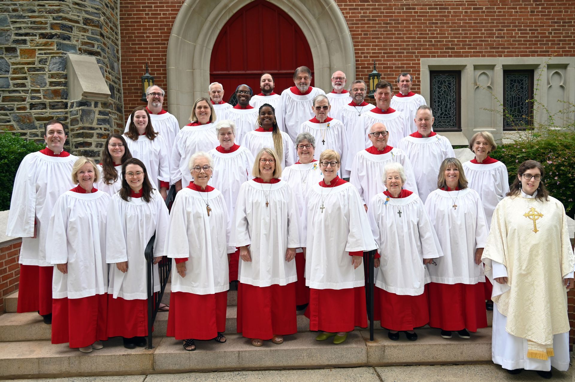 Choir group in white and red robes standing on church steps before a red arched door with a priest.