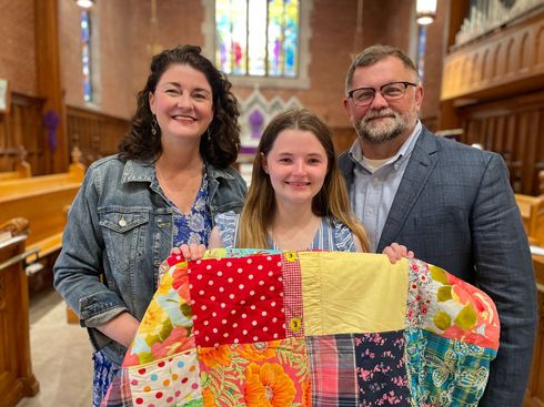 A family, daughter and parents, hold a colorful quilt inside a church sanctuary