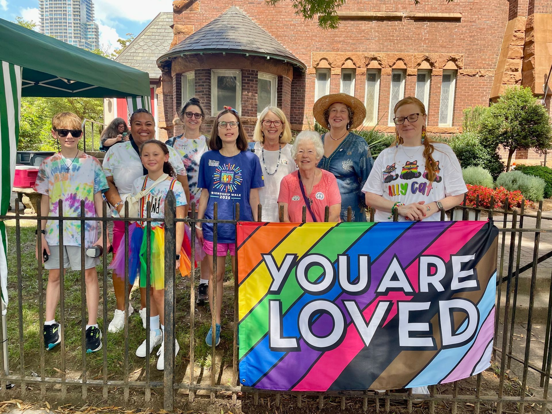 Group posing behind rainbow “YOU ARE LOVED” banner at a pride event outside a brick building