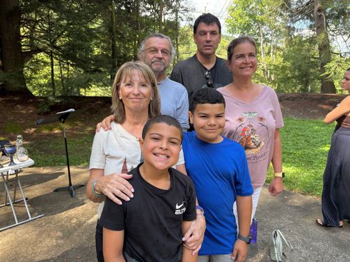Children and adults of all ages stand around a campfire roasting marshmallows at an outdoor gathering.
