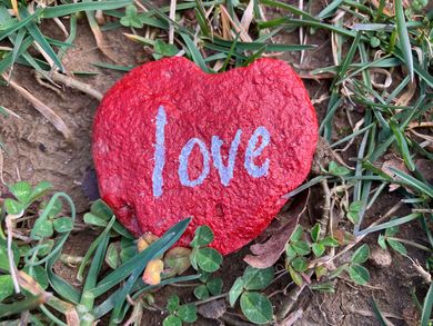 Rock in a heart shaped painted red with the word “love” in white, sitting on the grass.