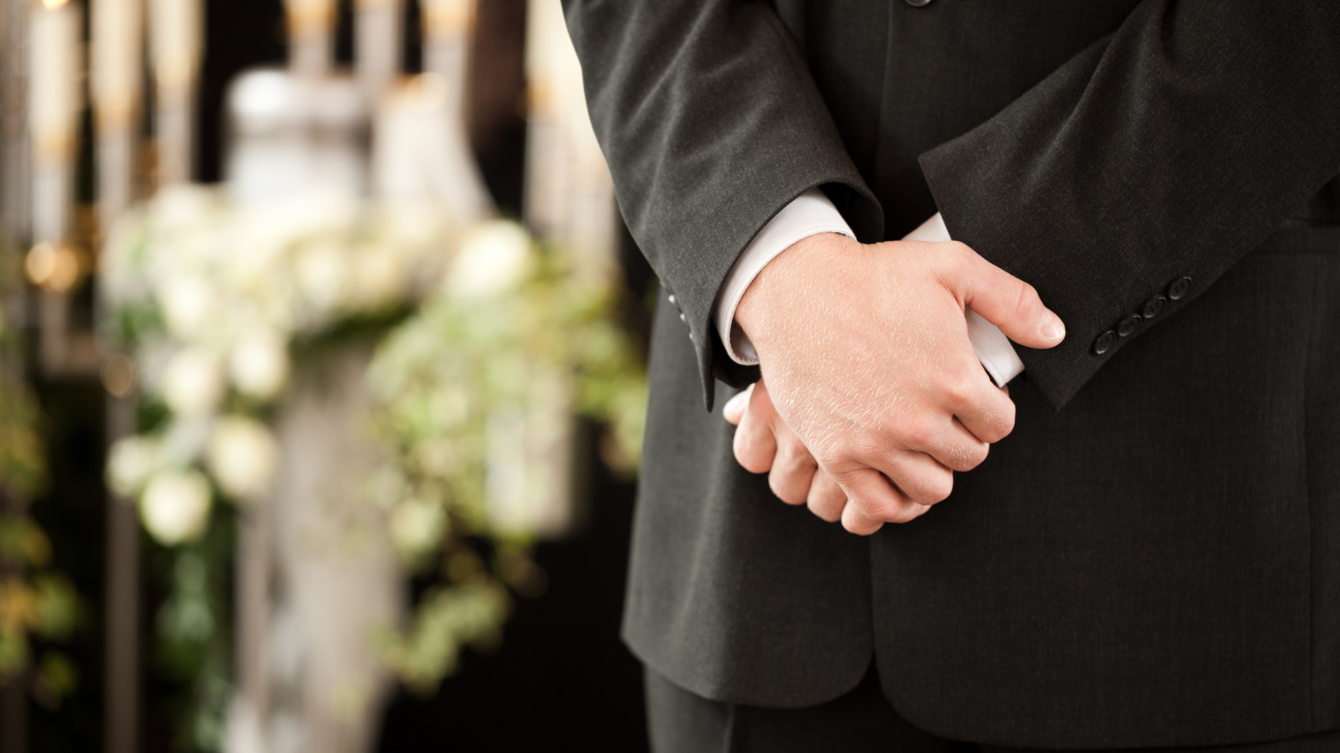 A man in a suit is standing with his hands folded at a funeral.