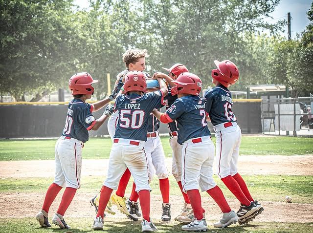 Youth baseball players in red helmets huddle and celebrate on a sunny field