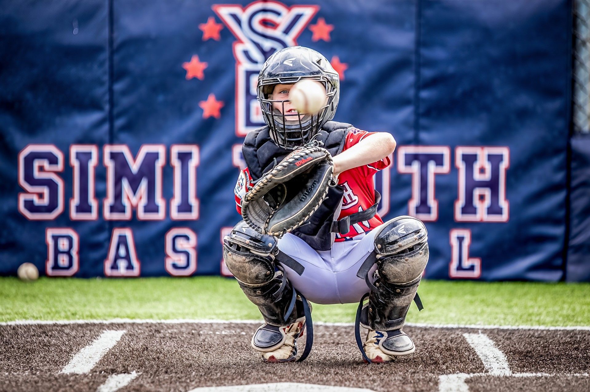 Youth baseball catcher crouching behind home plate in red-and-black gear at a Simi Valley field.