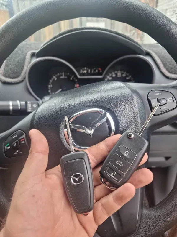 Hand Holding Two Car Keys in Front of A Mazda Steering Wheel — Nq Locksmiths In Kirwan, QLD