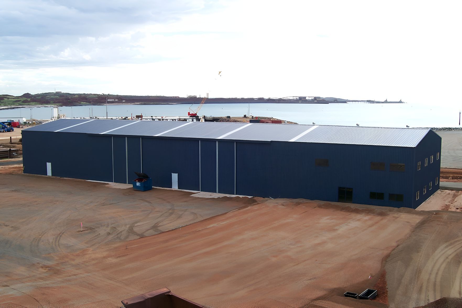 A Large Blue Building is Sitting in the Middle of a Dirt Field Next to a Body of Water — Mareeba Sheds & Gas In Mareeba, QLD
