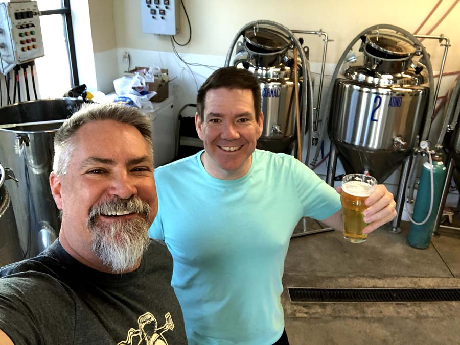 Two men are standing next to each other in a brewery holding glasses of beer.