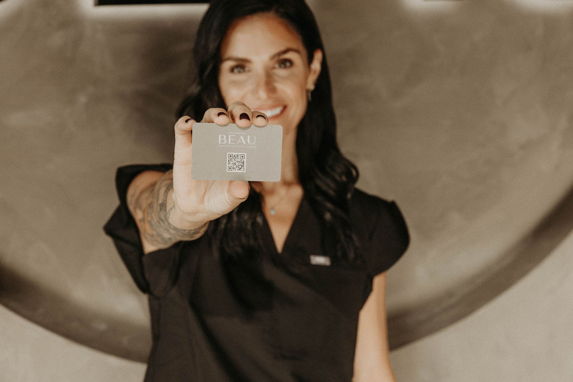 Woman smiling, holding a card with a QR code, wearing black scrubs.
