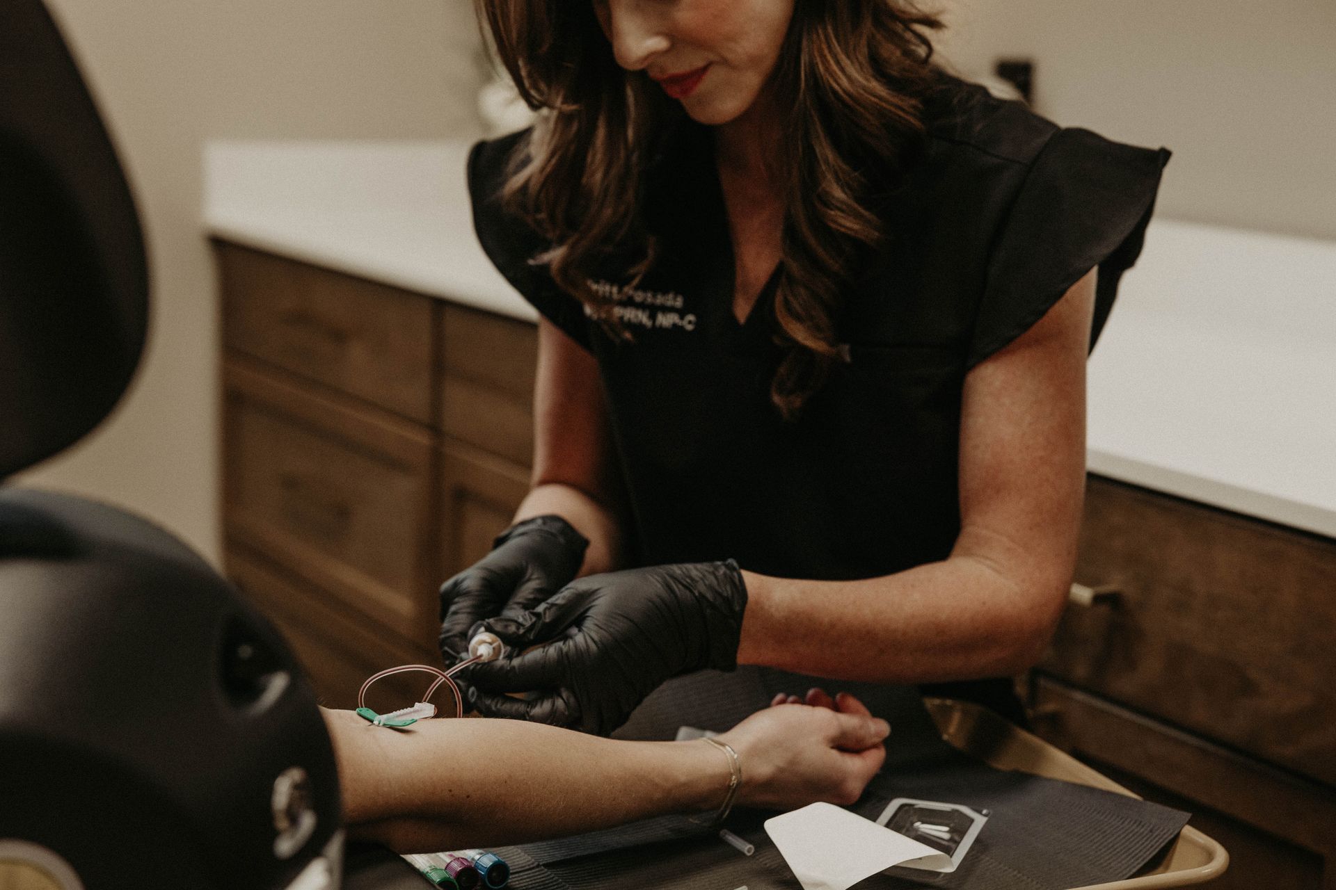 A woman in black scrubs, wearing gloves, administering an IV to a patient's arm.