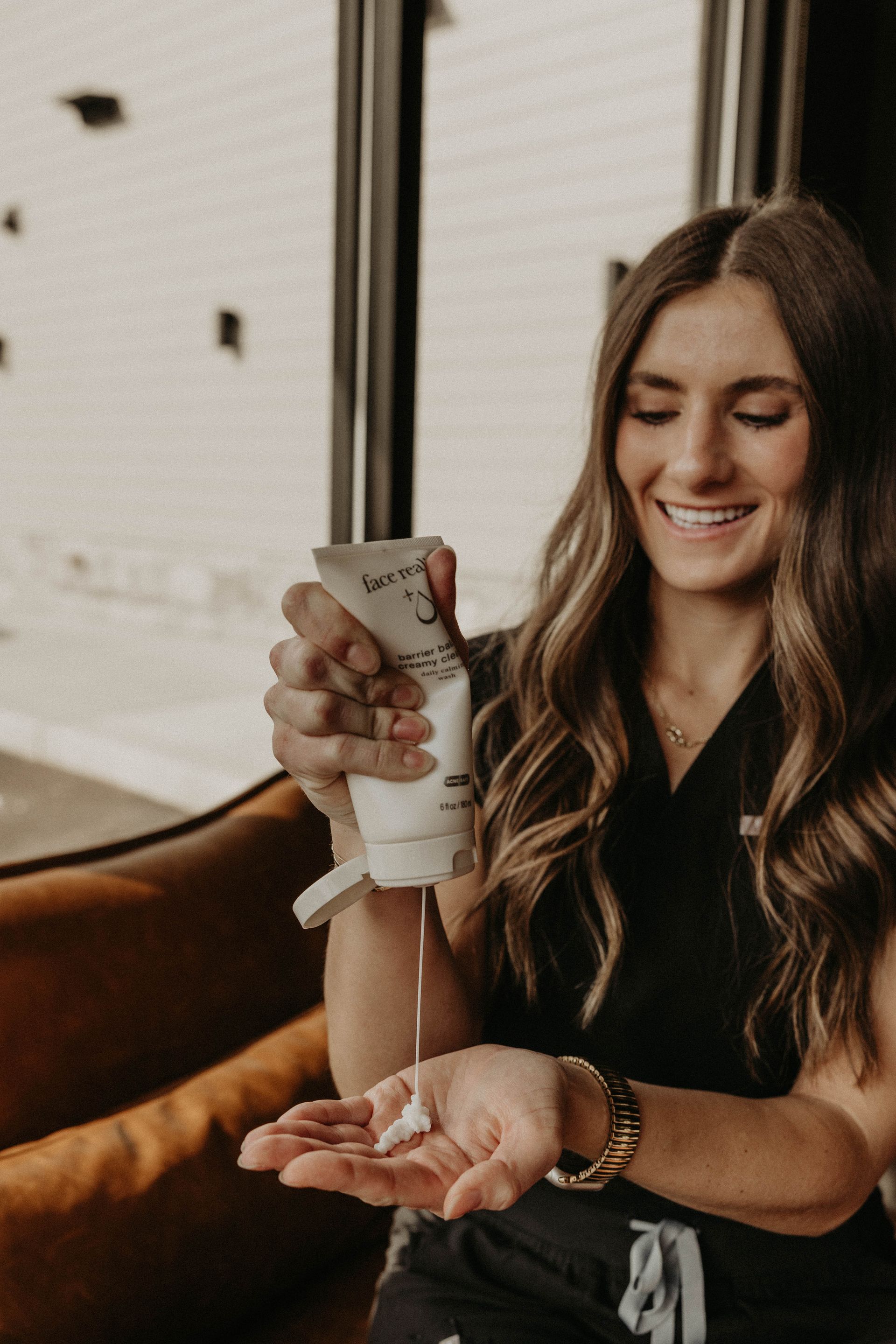 Woman pouring product from a tube into her hand, smiling; wearing scrubs, indoors.