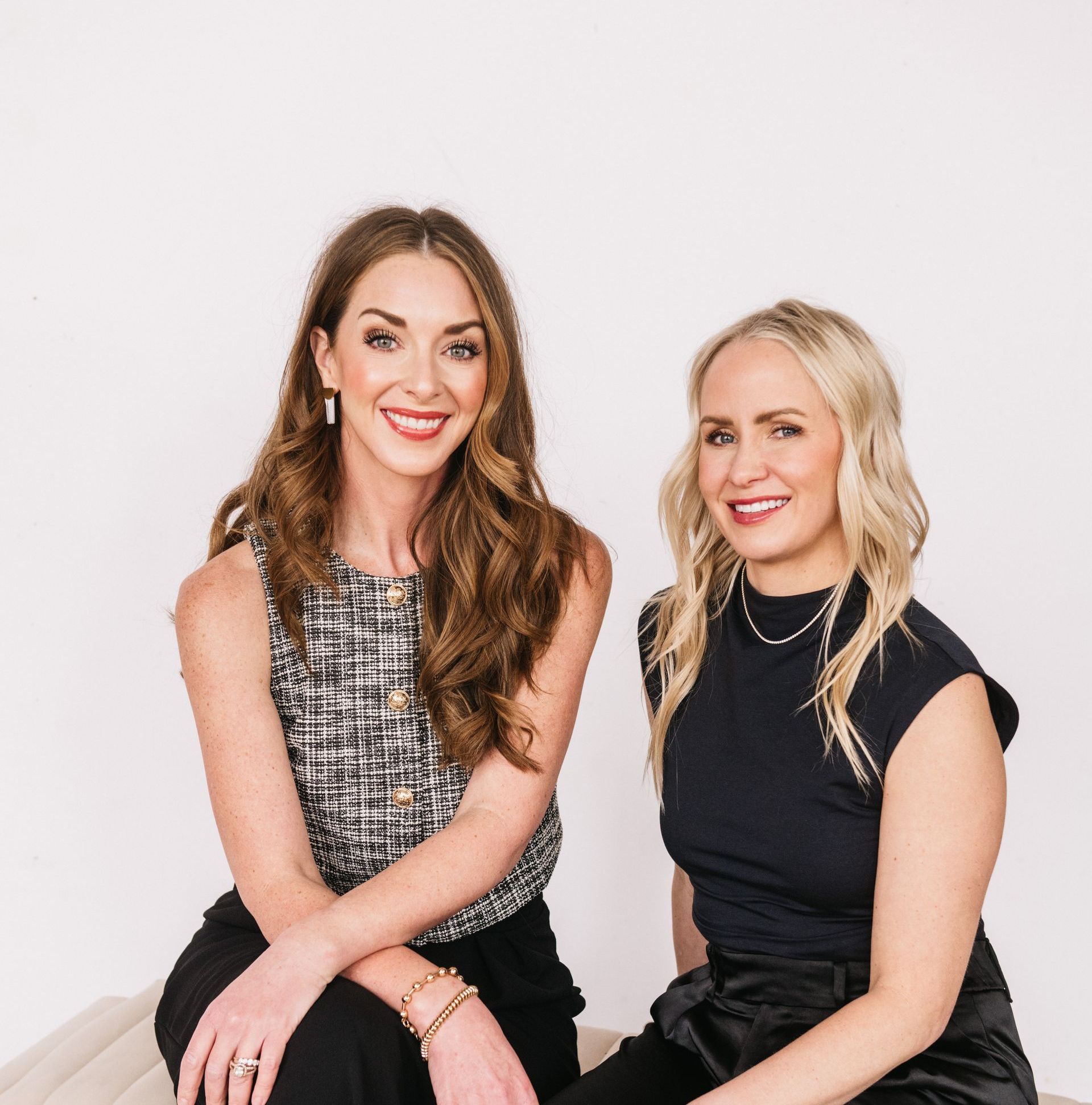 Two women smiling, seated. One wears a black and white patterned top, the other a black top. White backdrop.