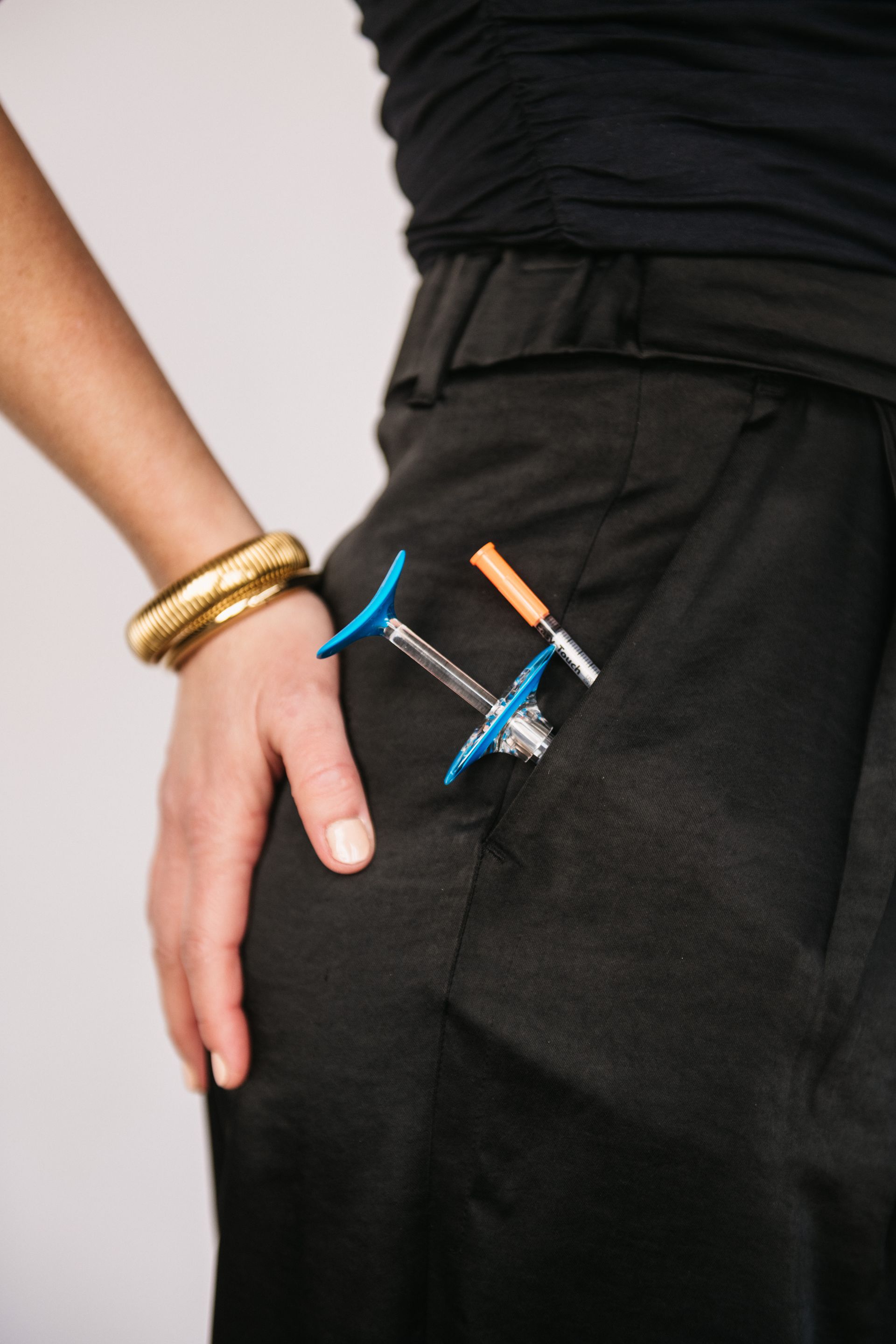 Woman's hand in a black pants pocket, holding a blue and clear medical device and a corkscrew, gold bracelet on wrist.