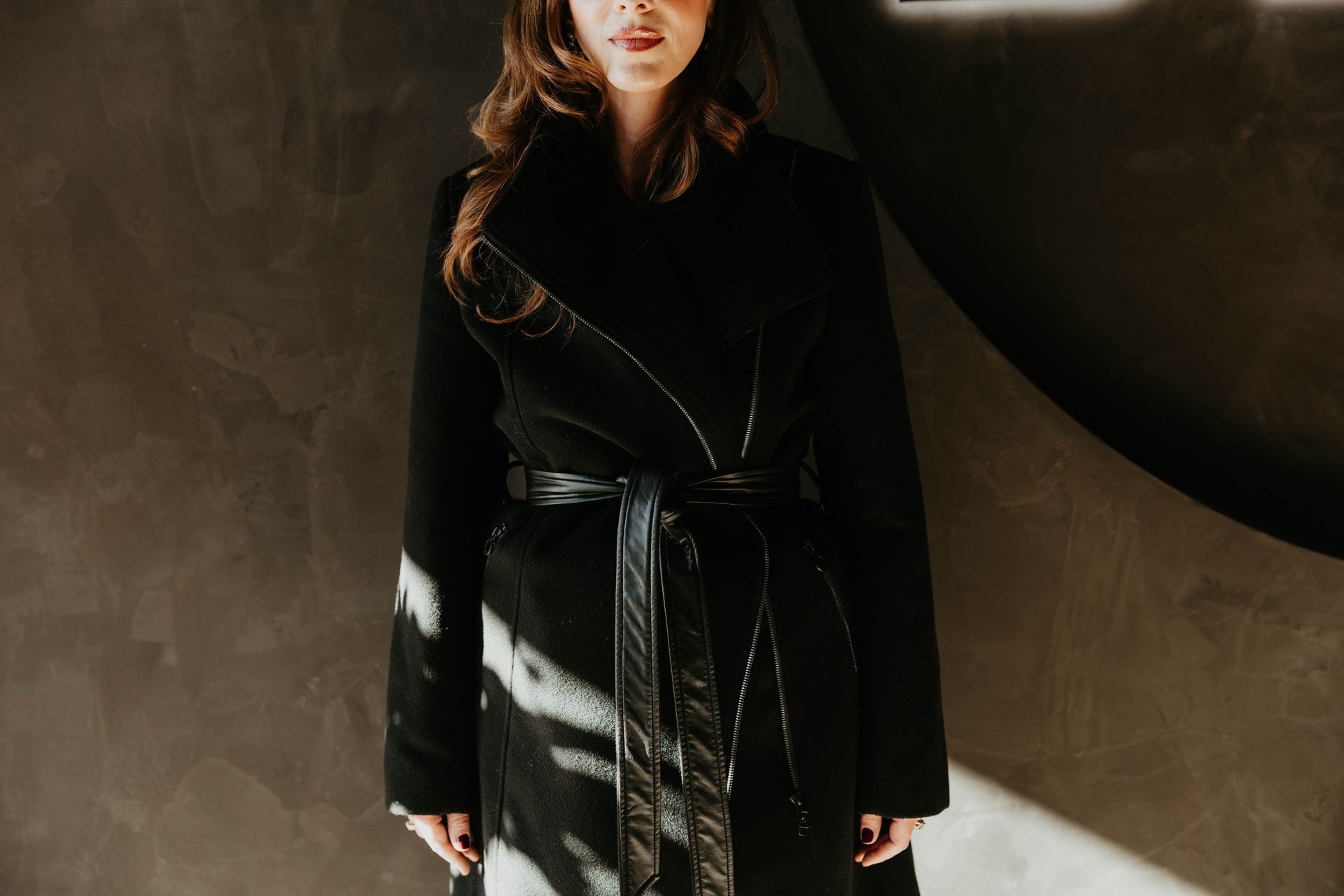 Woman in black coat with leather belt standing in front of a dark wall.