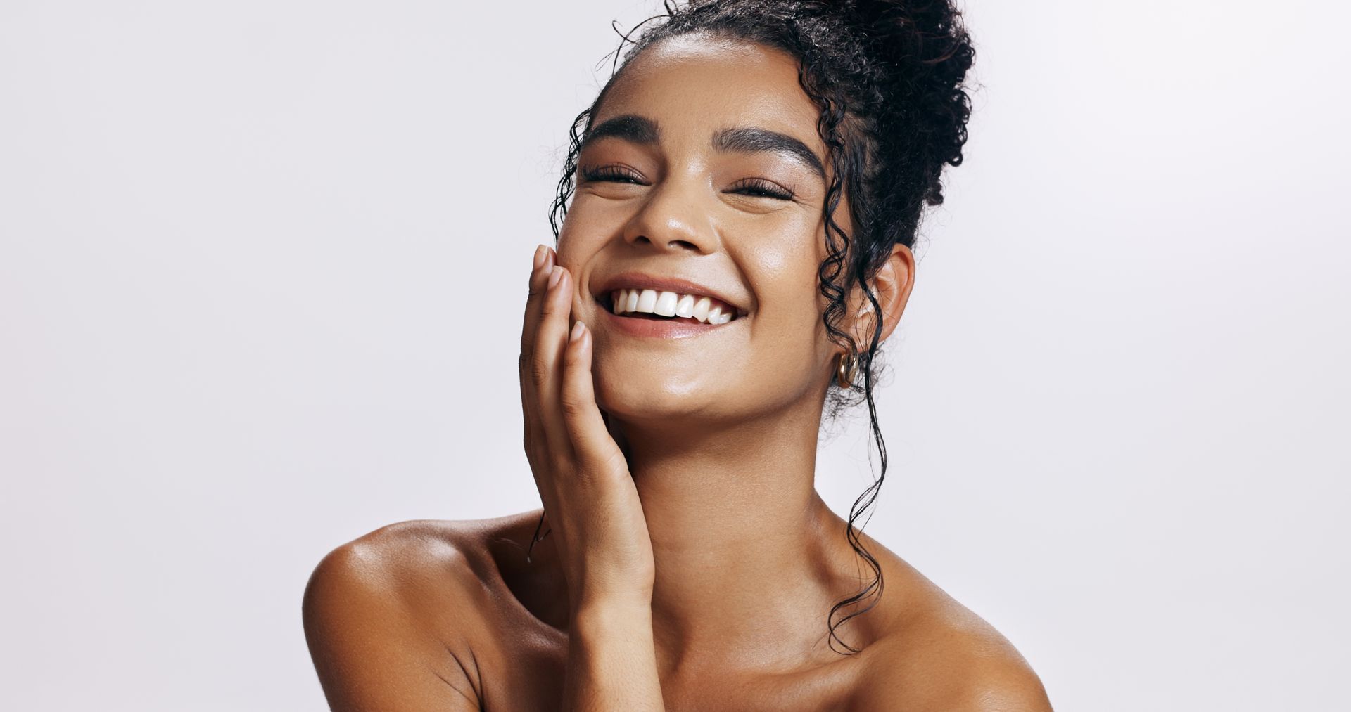 Smiling person with curly hair touching their face against a light background