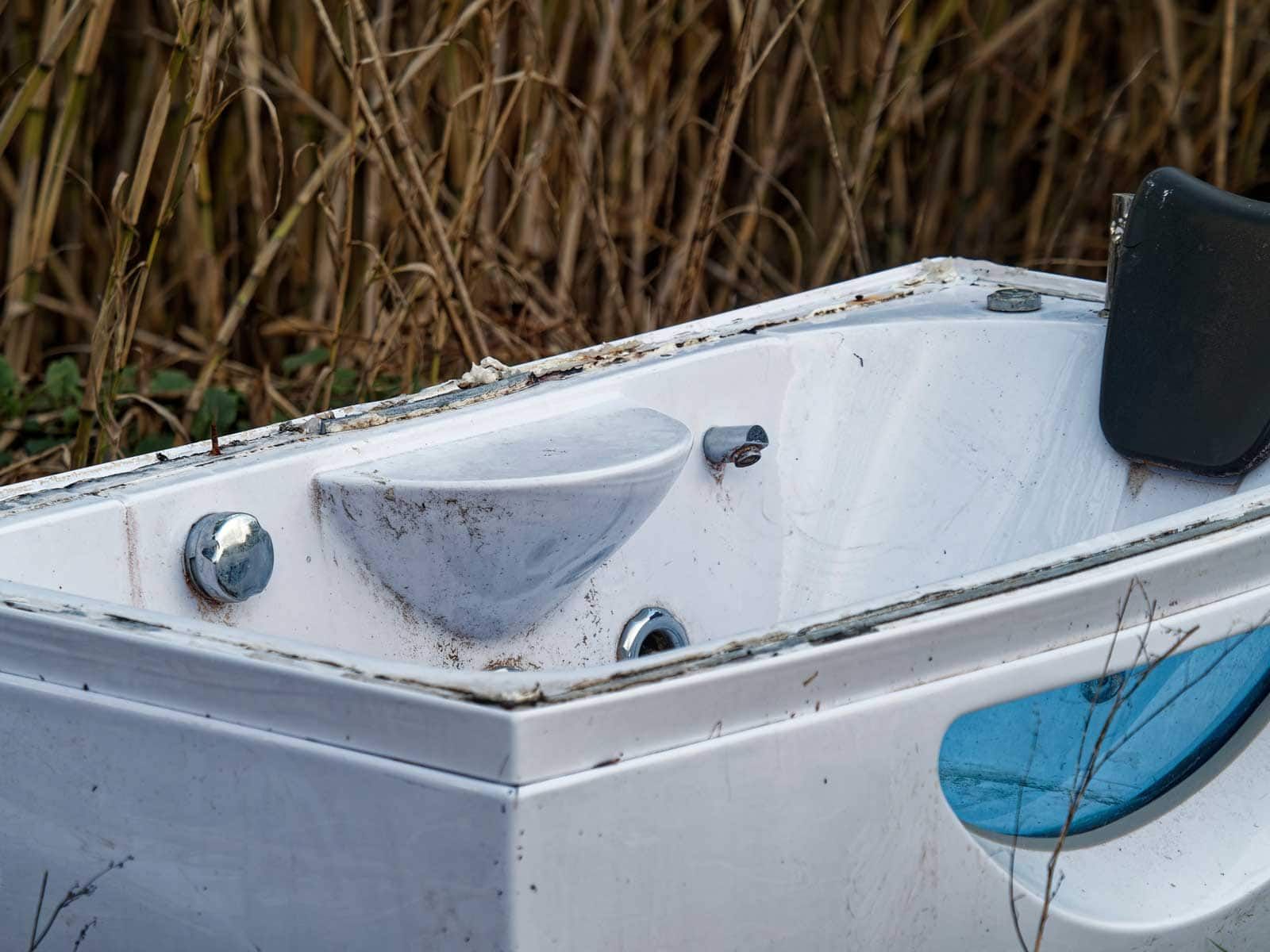 White, abandoned jacuzzi with blue accents, outdoors with brown foliage in the background.
