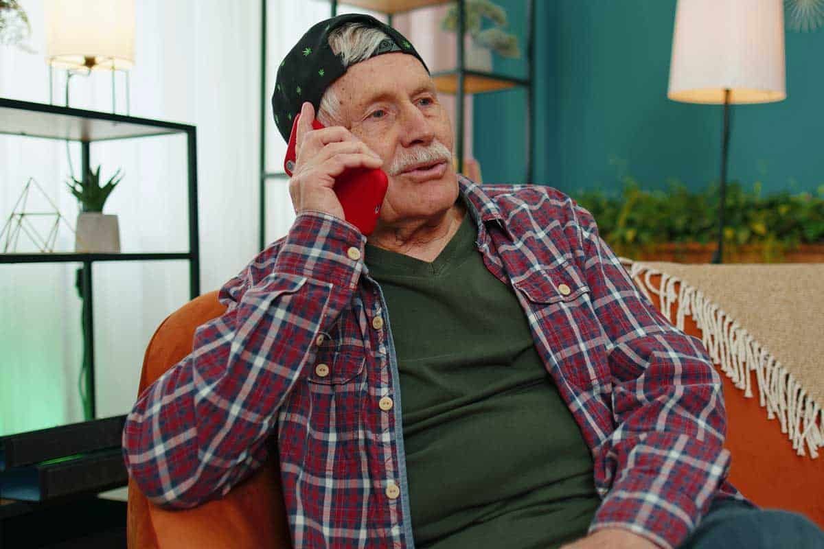 Elderly person in plaid shirt talking on red phone, sitting on orange couch indoors.