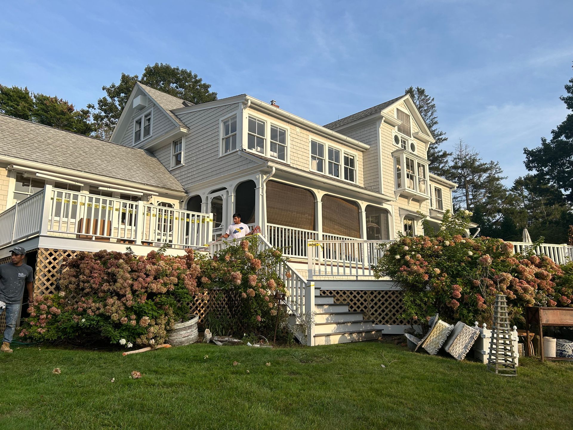 Large gray beach house with wraparound porch, white trim, and flowering shrubs on a sunny day