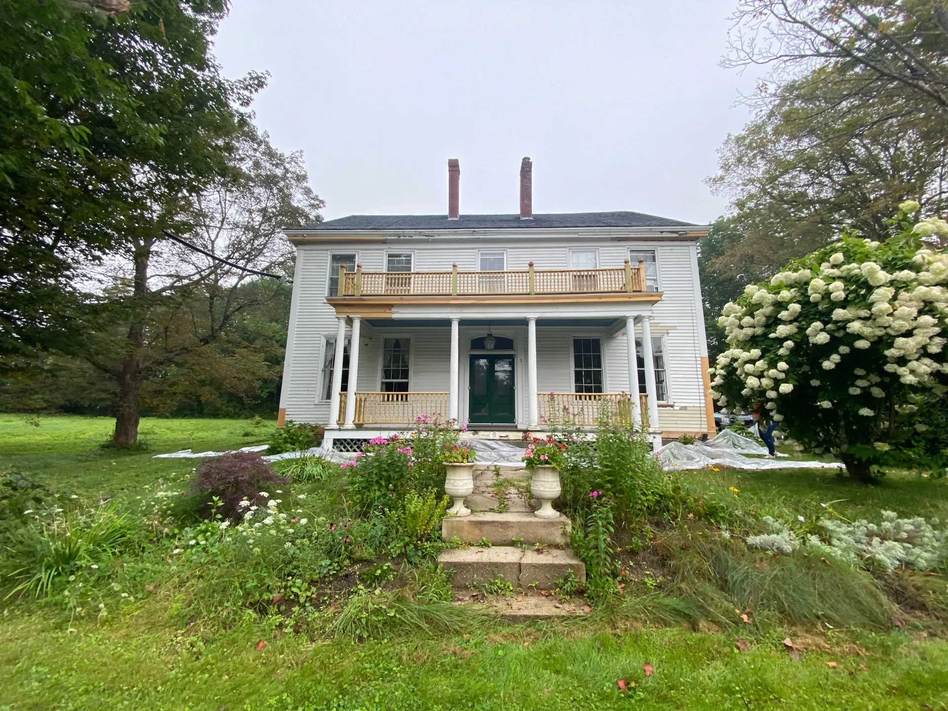 A large white house with a large porch is surrounded by trees and flowers.