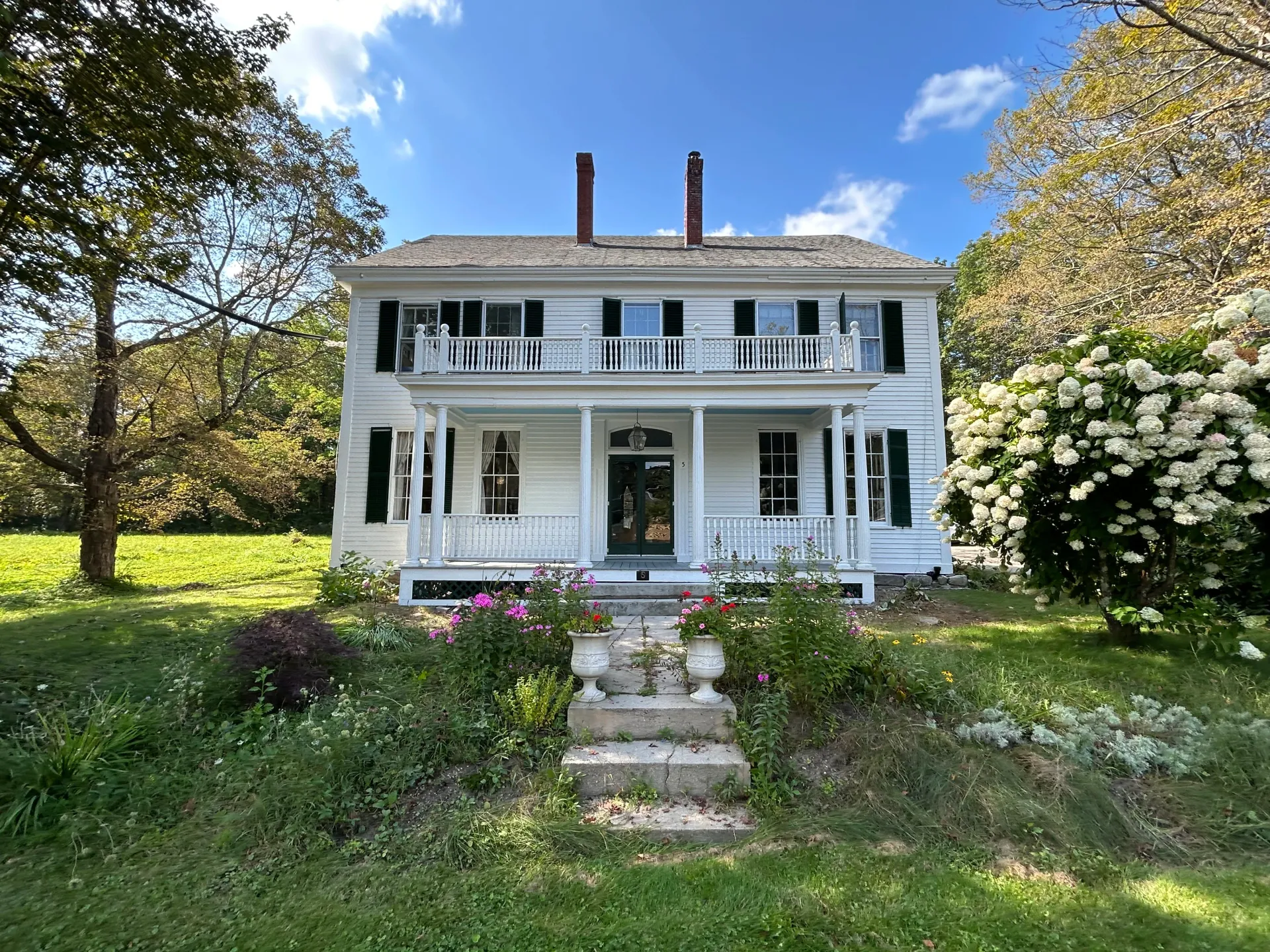 A large white house with a large porch is surrounded by trees and flowers.