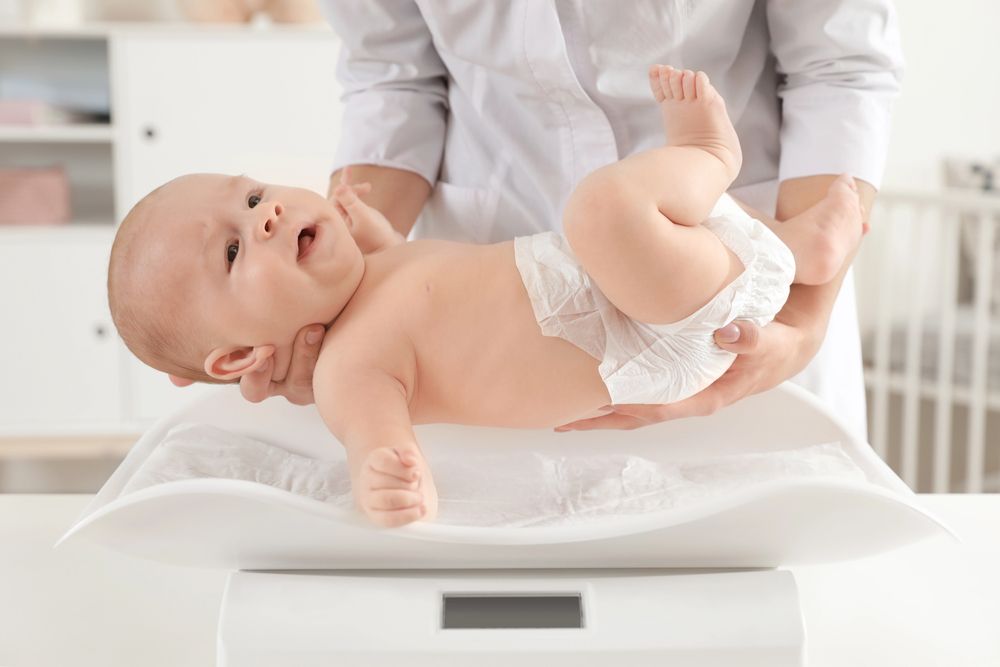 Un neonato su una bilancia che cambia colore, con una persona che gli tiene le gambe sollevate in una stanza luminosa.