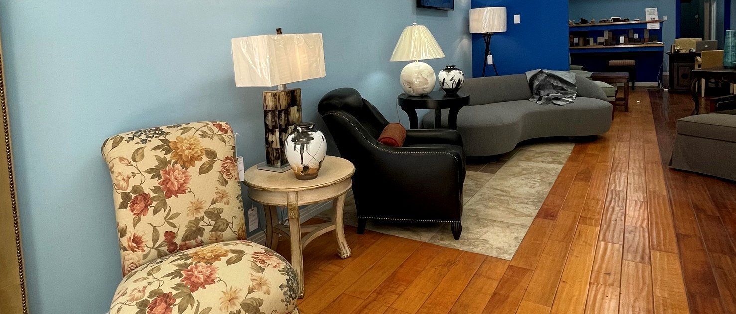 A living room with a floral chair, black leather recliner, grey curved couch, lamps, and wooden floor.