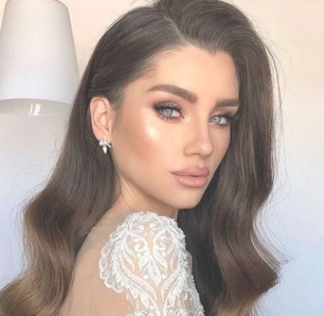 Donna con capelli castani ondulati, trucco glamour e abito bianco di pizzo.
