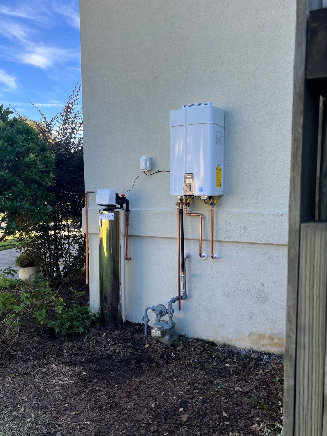 Exterior wall with a white water heater, pipes, and utility meters beside a house.