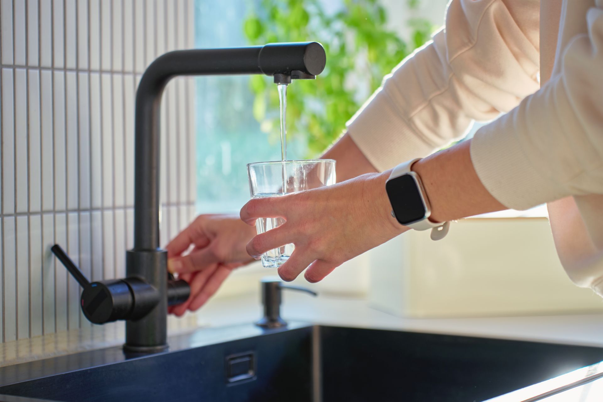 Hands filling a glass with water from a black kitchen faucet at a sink