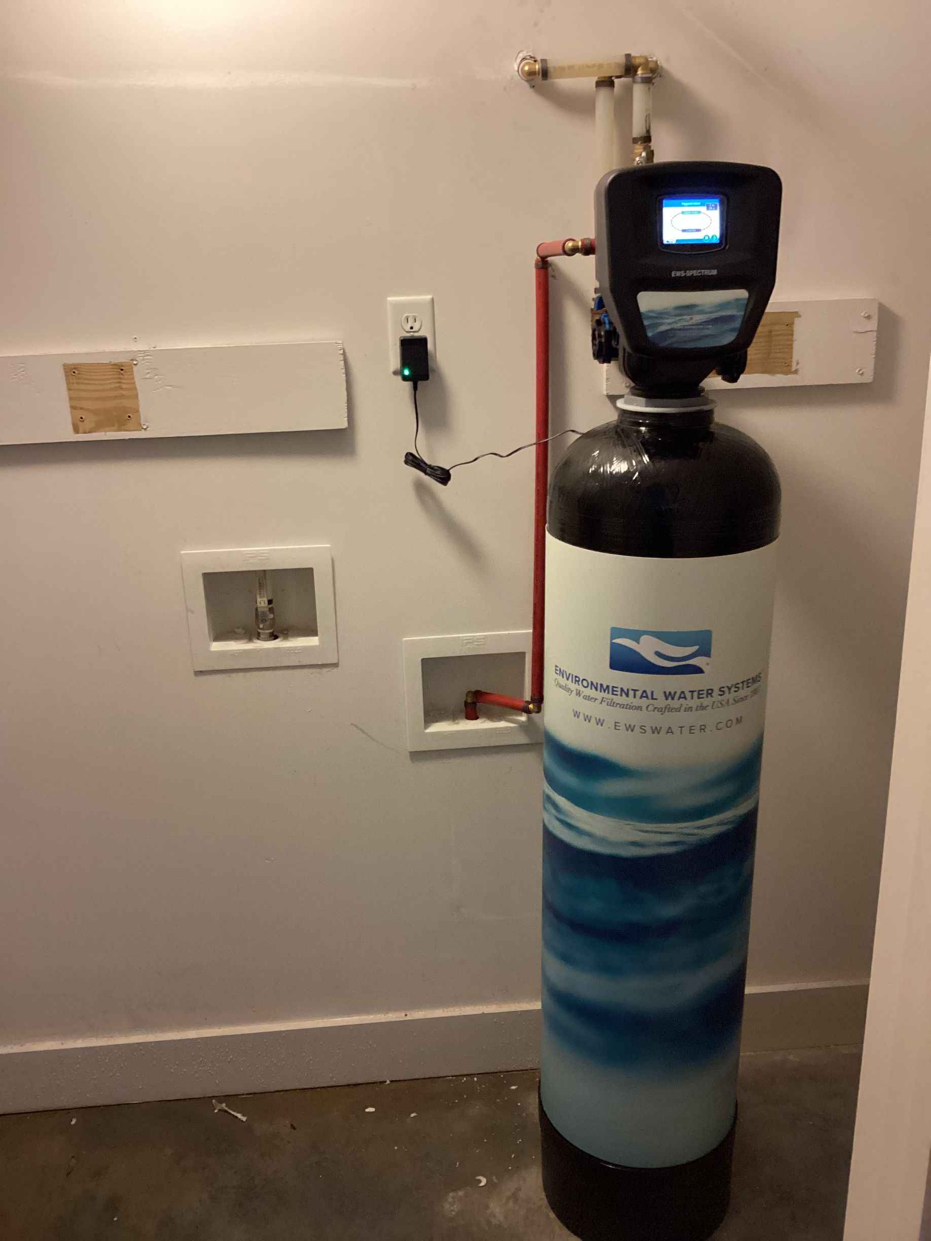 Water softener tank with digital control in a utility room, white wall and exposed plumbing behind it
