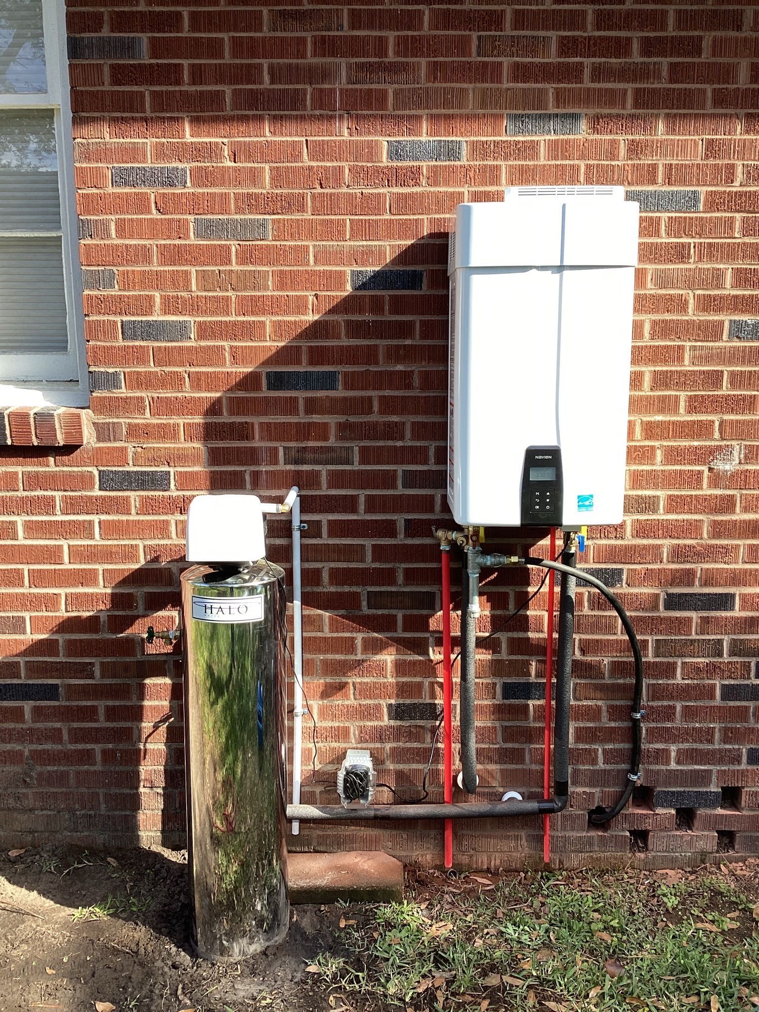 Outdoor tankless water heater mounted on a brick wall with exposed red and black pipes beside a utility post.