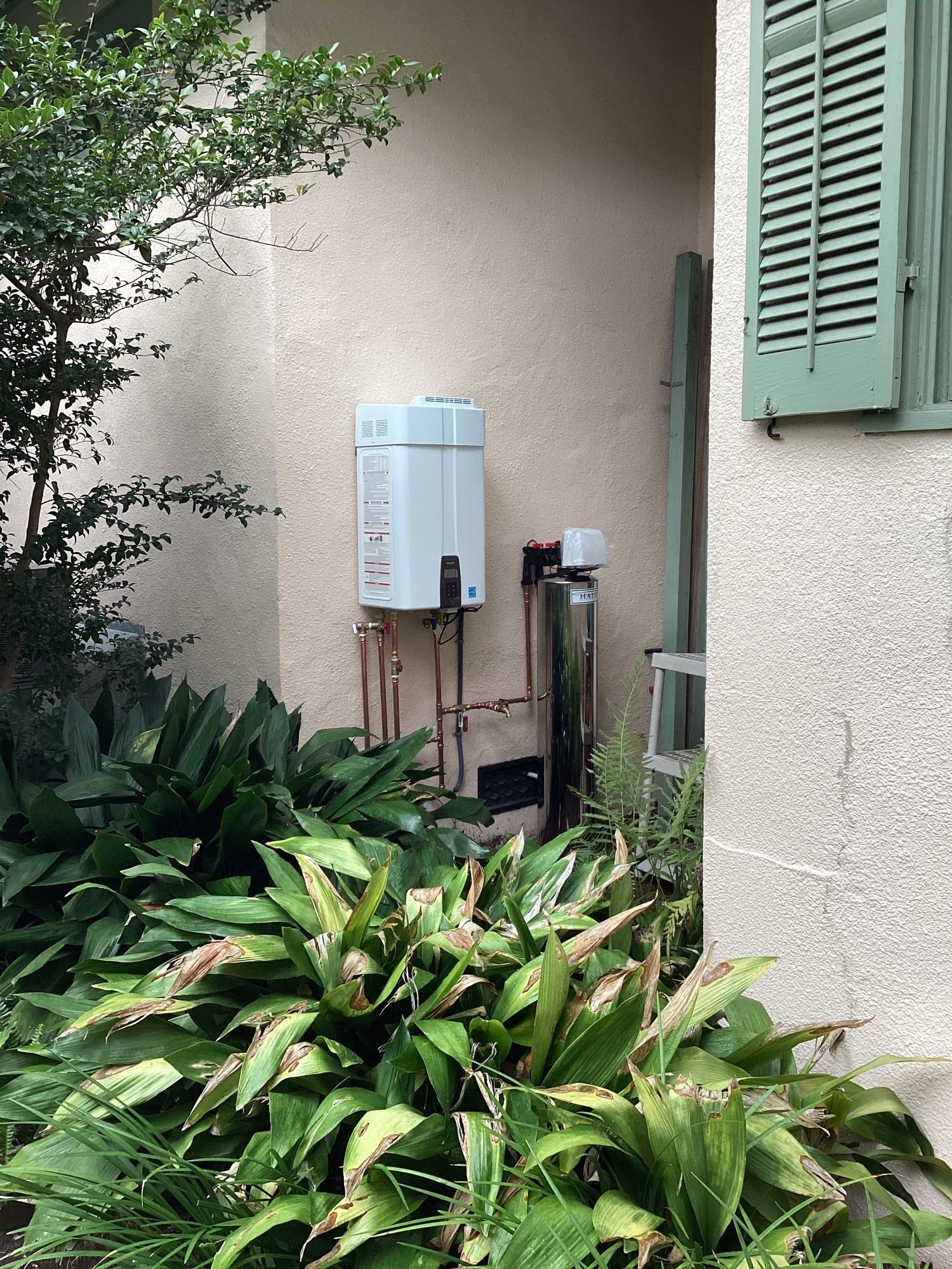 Outdoor utility meter and boxes beside a beige house, partly hidden by green plants and a green shutter.