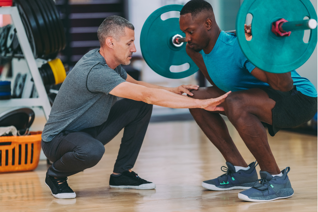 Man lifting weights with trainer in a gym; trainer guiding his form.