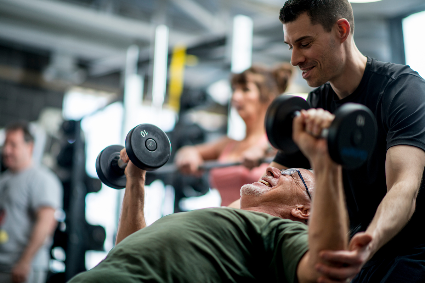 Man lifting dumbbells with trainer's assistance at a gym; other people exercise in background.