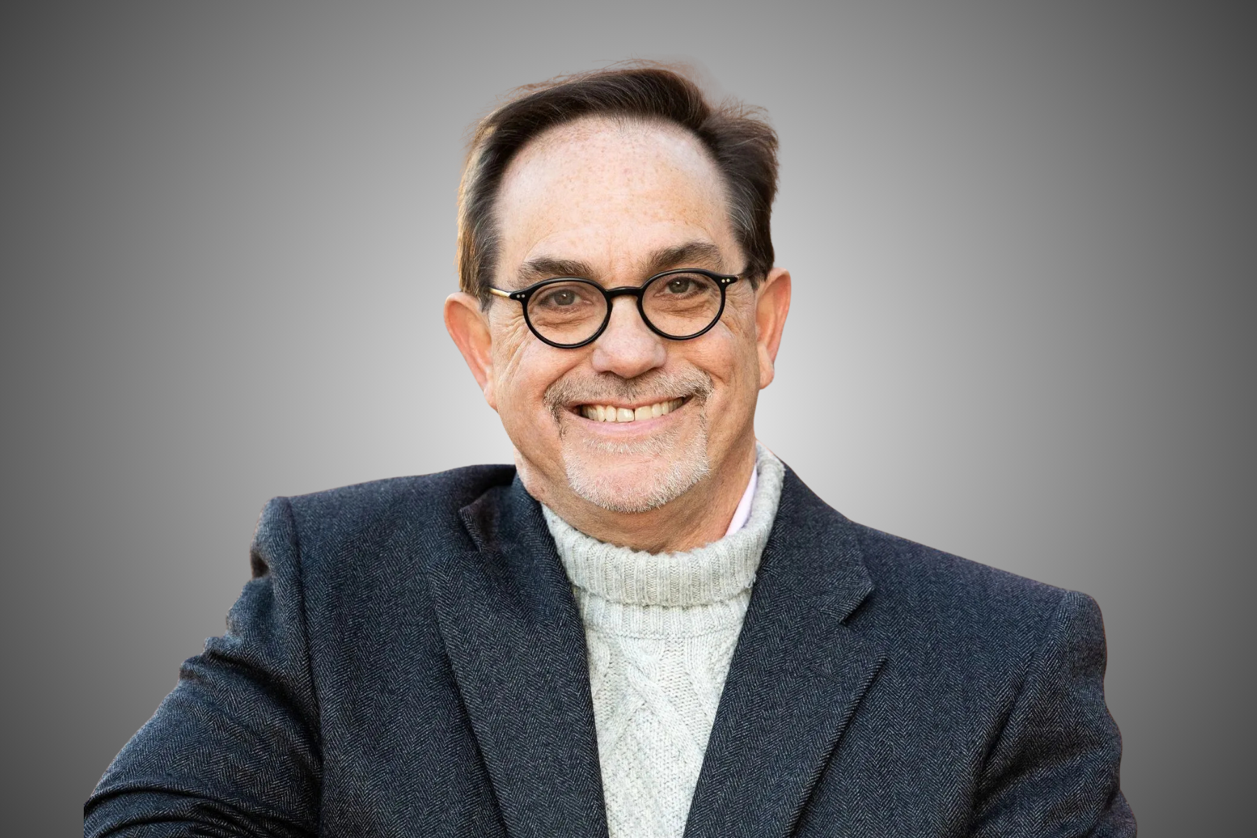 Man with glasses smiles, wearing a navy jacket and white sweater. Gray background.