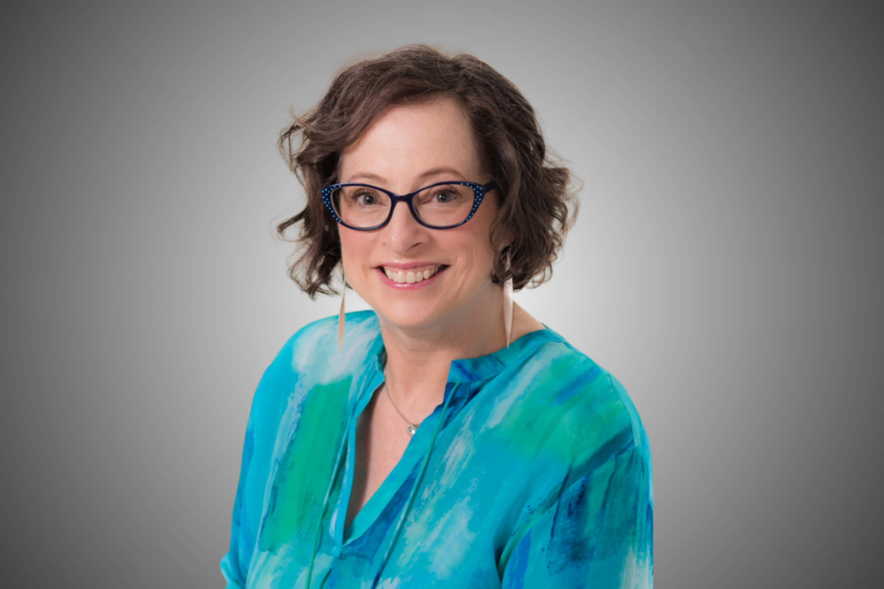 Woman with glasses smiles, wearing blue patterned top, against a gray gradient background.