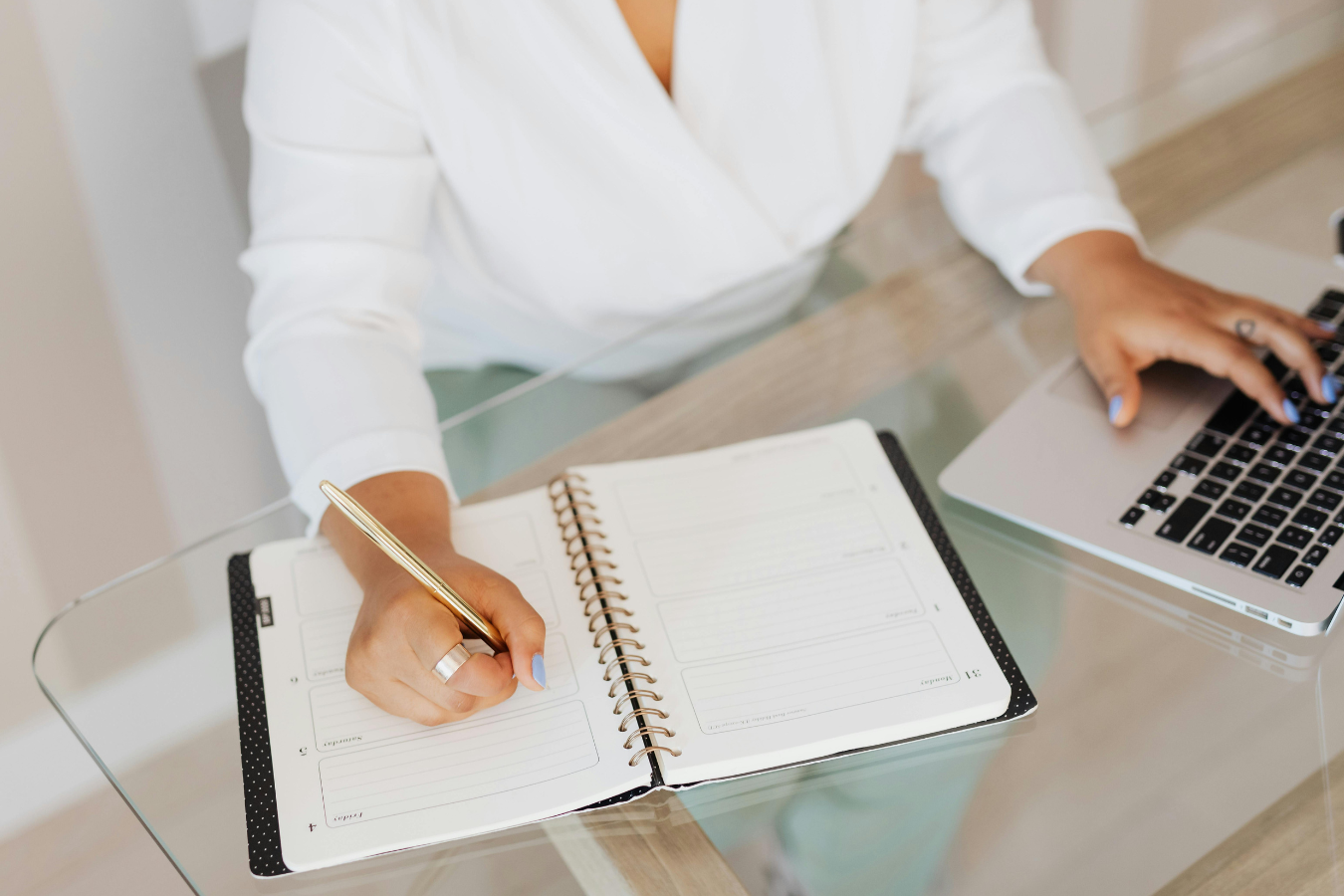Woman in white top writing in a notebook next to a laptop on a glass desk.
