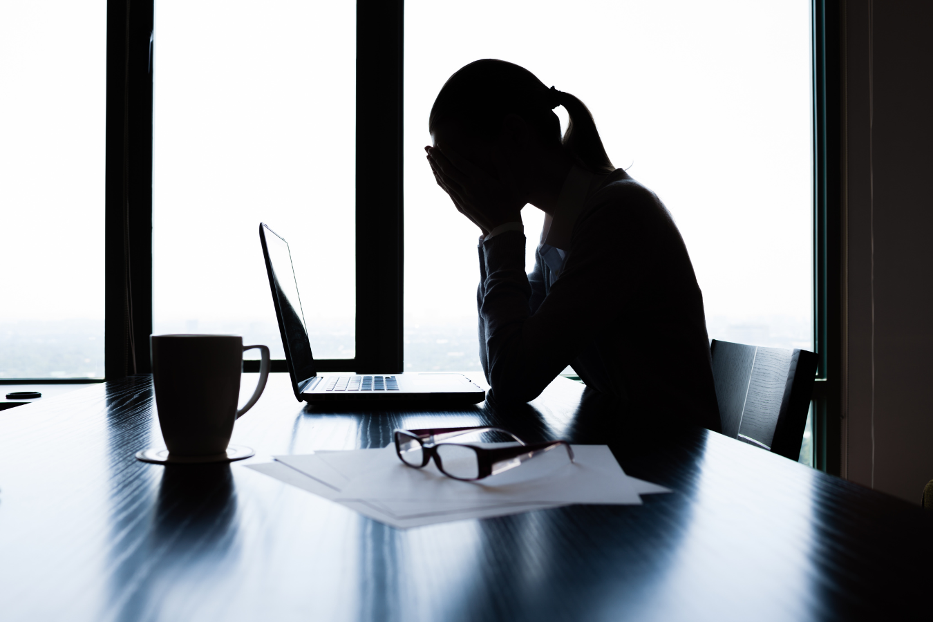 Silhouette of a person at a desk with head in hands, laptop, coffee mug, and papers visible.