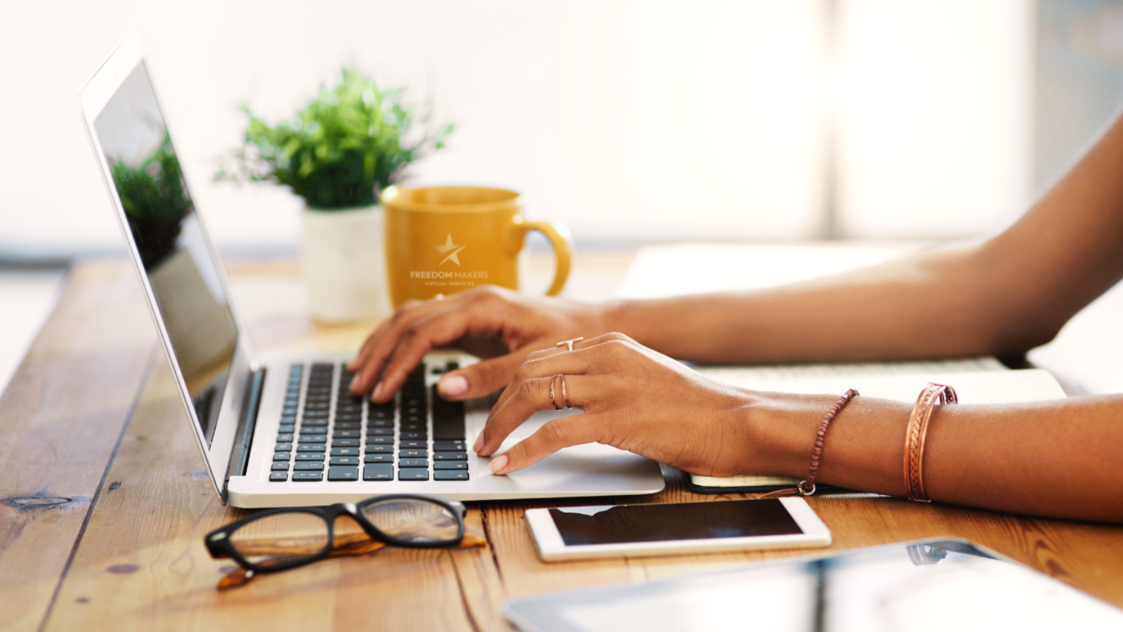 Hands typing on a laptop at a wooden desk, mug, glasses, and tablet present.