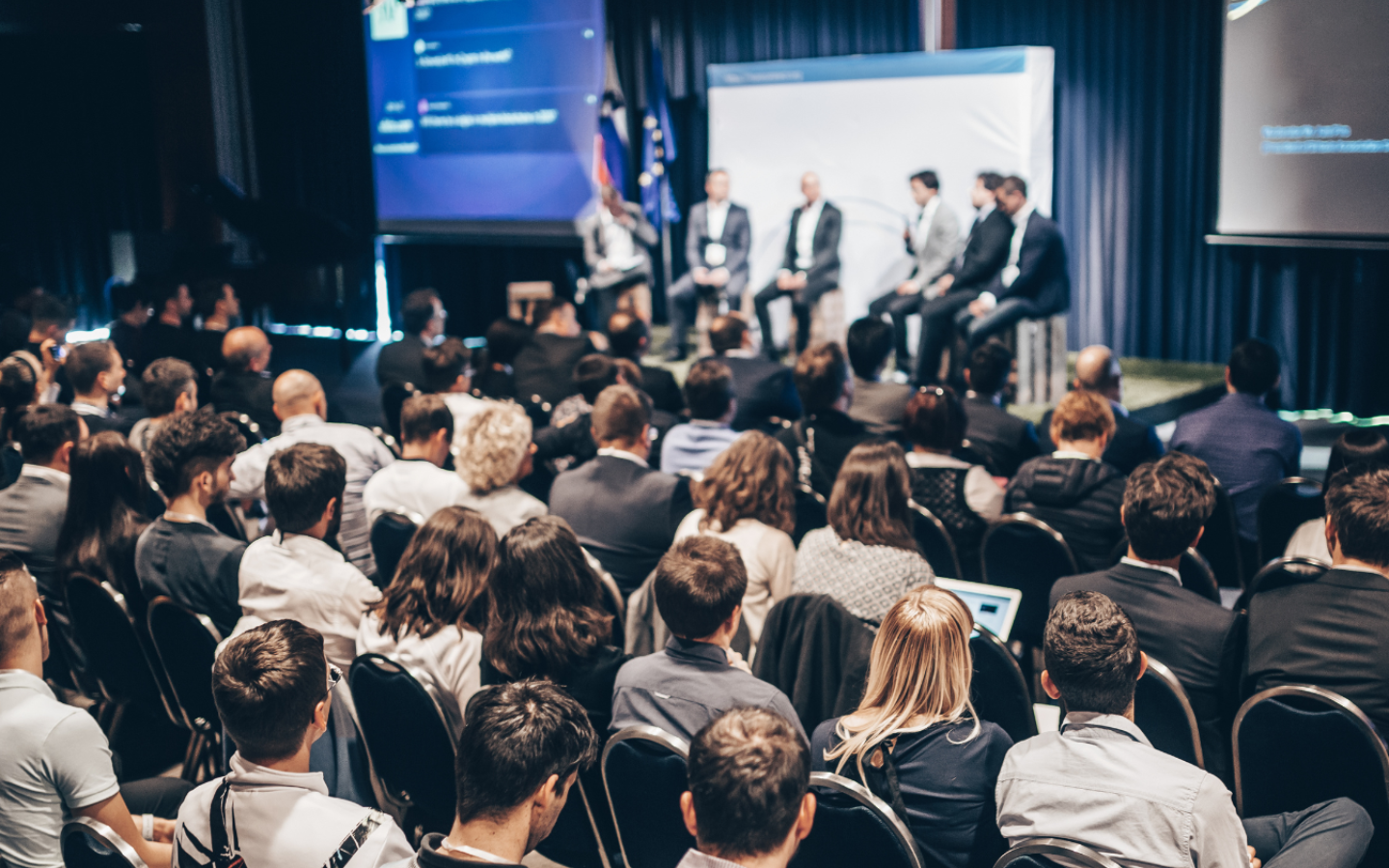 Audience at a conference watches a panel discussion on stage with five speakers and two screens.