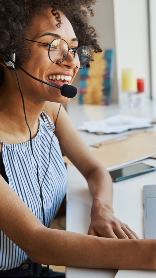 Woman wearing headset, smiling, working on laptop at a desk.