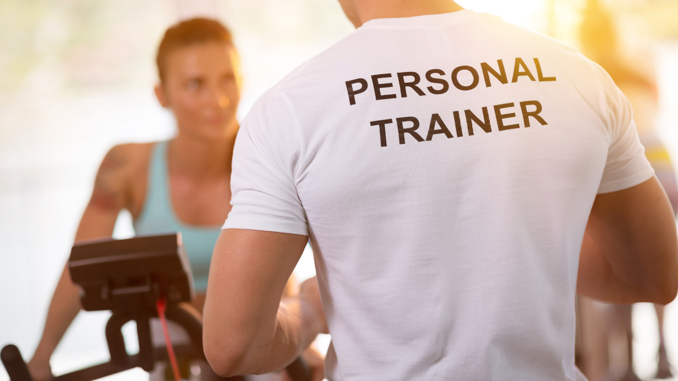 a man wearing a personal trainer shirt is talking to a woman on an exercise bike
