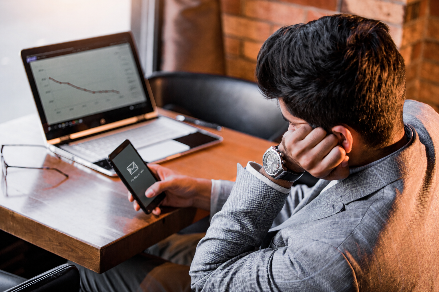 Person sitting at a table with a laptop and smartphone, looking at a stock chart on the laptop screen.