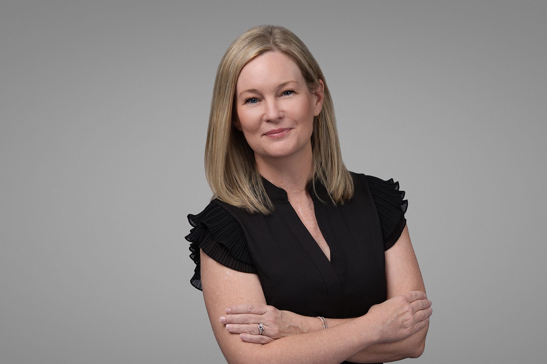 Woman with blonde hair, wearing a black shirt, arms crossed, smiling. Gray background.