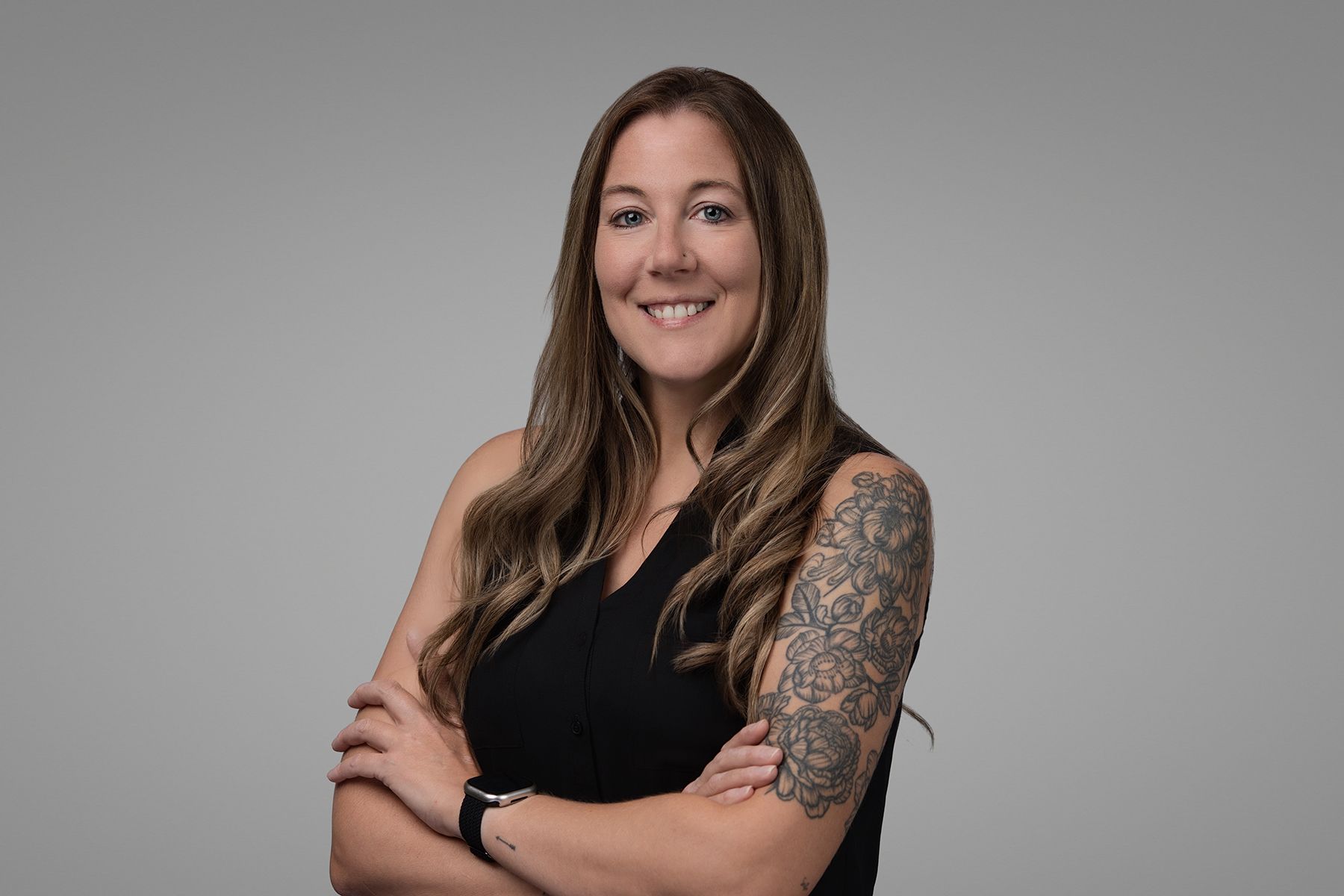 Woman with long brown hair, arm tattoos, black dress, arms crossed, smiling.