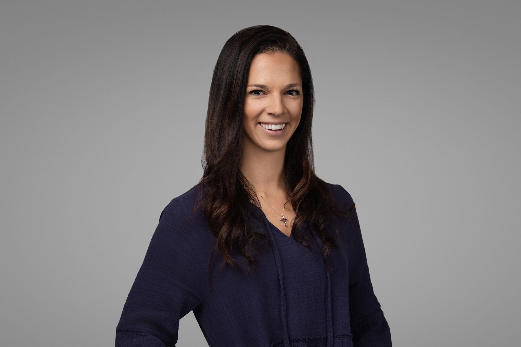 Woman smiling, dark hair, blue top, hands at sides, against a gray background.