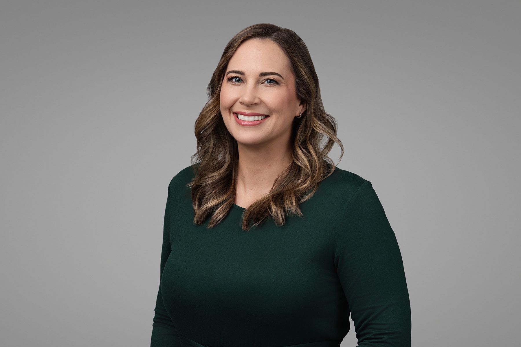 Woman in green long-sleeve shirt smiling, looking at the camera, against a gray background.