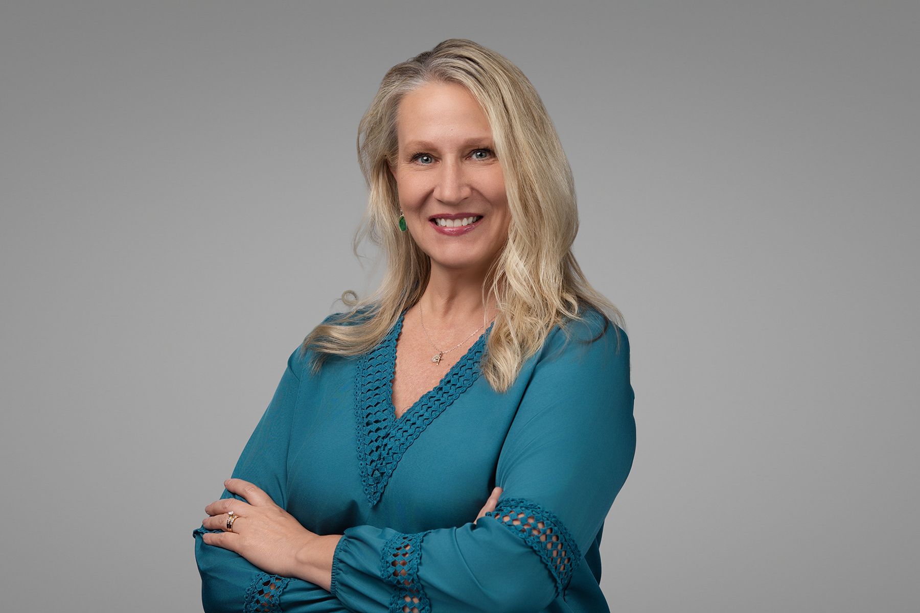 Woman with blonde hair wearing a teal blouse smiles with arms crossed. Gray background.
